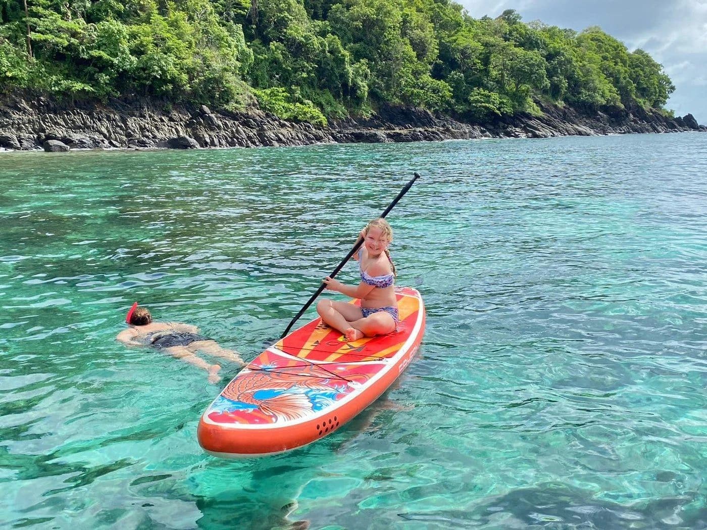 Girl paddling on a stand-up board beside a snorkeler in turquoise water